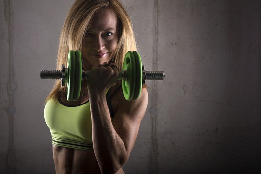 A fit, athletic blonde woman wearing a sports bra, smiles as she exercises with a green dumbbell. Her flexed muscles show toned abdominals, triceps and biceps.