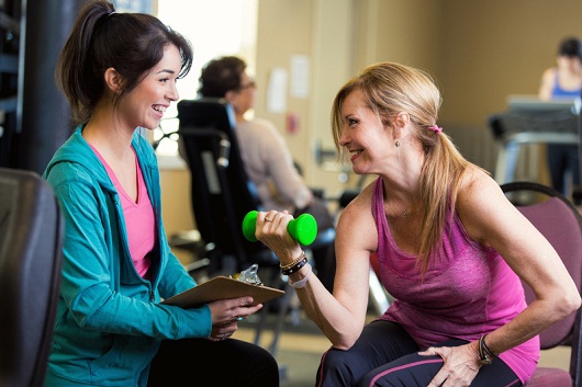 Caucasian female personal trainer working with senior client at community center. The woman is lifting weights as the trainer coaches her.