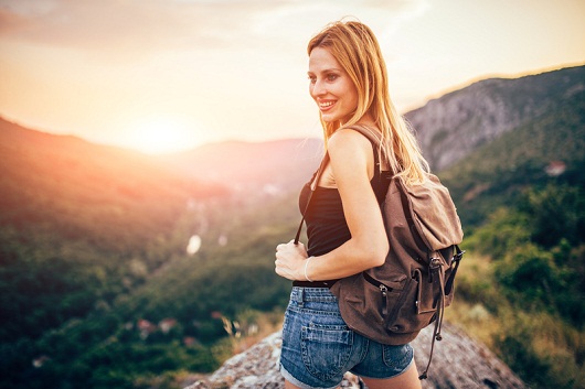 Young lady hiker standing with backpack on top of a mountain and enjoying a sunny summer day